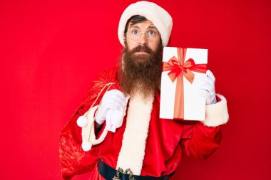 Uzun sakallı yakışıklı kızıl saçlı, Noel Baba kostümü giyen ve hediyeli büyük çantalı, yüzünde ciddi bir ifadeyle rahatlamış bir adam. Basit ve doğal bir şekilde kameraya bakıyorsun.. 
