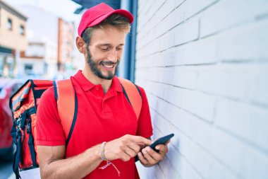 Beyaz teslimatçı kırmızı üniforma giyiyor ve sırt çantası teslim ederken gülümsüyor ve akıllı telefon kullanıyor.