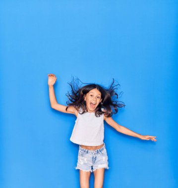 Sevimli İspanyol kız günlük kıyafetler giyip mutlu gülümsüyor. İzole edilmiş mavi arka planda yüzünde gülümsemeyle zıplıyor