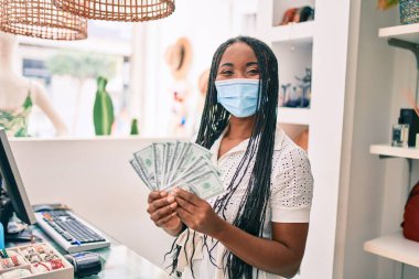 Tıbbi maske takan genç Afro-Amerikalı kadın giyim mağazasında dolar banknotları tutuyor.