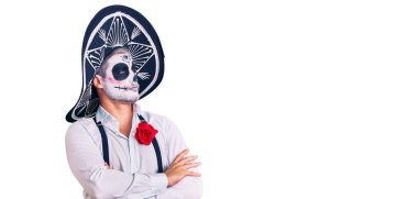 Arka planda ölü kostümü giymiş bir adam kollarını kavuşturmuş ve kendinden emin bir şekilde yan tarafa bakıyor. 