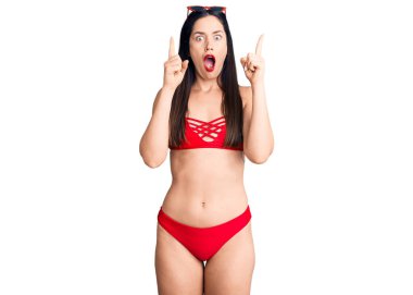 Bikini giyen güzel beyaz kadın şaşırmış ve şaşırmış bir şekilde yukarı bakıyor ve parmaklarıyla işaret ediyor ve kollarını kaldırıyor.. 