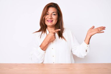 Orta yaşlı, esmer, Latin iş kadını. Masada oturan, hayrete düşmüş, kameraya gülümseyen, eli ve parmağı olan bir kadın.. 