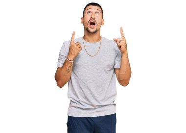 90 'ların tarzında dövmeleri olan yakışıklı bir adam şaşırmış ve şaşırmış bir şekilde yukarı bakıyor ve parmaklarıyla işaret ediyor ve kollarını kaldırıyor.. 
