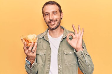 Patates cipsi tutan yakışıklı genç adam parmaklarıyla imza atıyor, gülümseyerek mükemmel bir sembol oluşturuyor. 