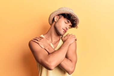 Genç İspanyol adam yaz şapkası takıyor mutlu ve olumlu bir şekilde kendine sarılıyor, kendinden emin gülümsüyor. Sevgi ve özgüven. 