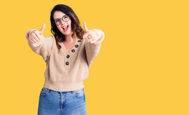 Güzel, genç, esmer bir kadın, günlük kıyafetler ve gözlük takıyor. Onaylıyor. Elleriyle olumlu bir jest yapıyor, gülümsüyor ve başarı için mutlu. kazanan jest. 