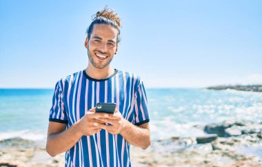 Genç İspanyol adam sahilde akıllı telefon kullanarak mutlu bir şekilde gülümsüyor..