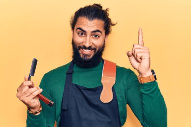 Önlük takan genç Arap adam elinde berber jiletiyle gülümsüyor bir fikir ya da soruyla gülümsüyor ve mutlu bir yüzle işaret ediyor. 