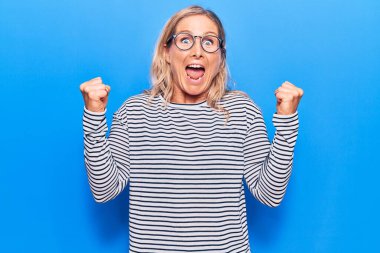 Orta yaşlı, beyaz, çizgili süveter ve gözlük giyen sarışın kadın kolları açık ve kolları açık bir şekilde başarıyı kutluyor ve şaşırıyor. Kazanan konsept. 