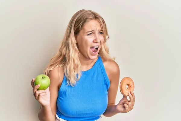 Chica Rubia Joven Sosteniendo Manzana Verde Donut Enojado Loco Gritando —  Fotos de Stock