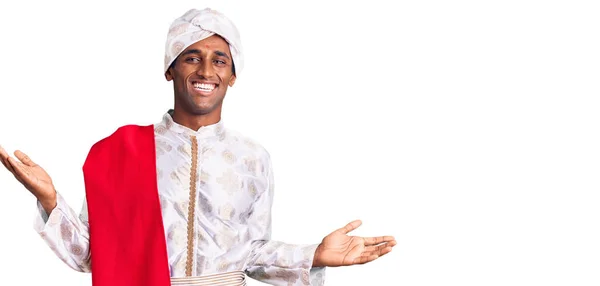 伝統的なシャーワニを身に着けているアフリカのハンサムな男は両手を開いてヤシの木を示す笑顔服を見て 提示し 広告の比較とバランス — ストック写真