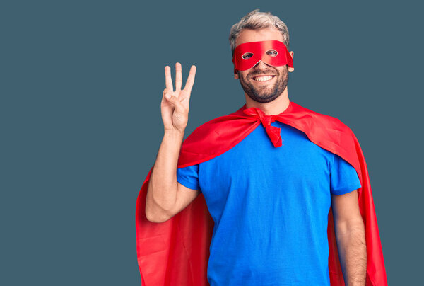 Young blond man wearing super hero custome showing and pointing up with fingers number three while smiling confident and happy. 
