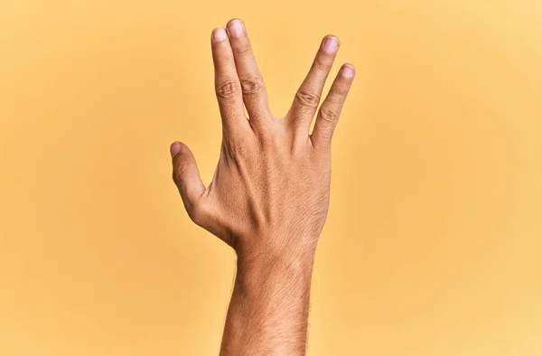 Arm and hand of caucasian man over yellow isolated background greeting doing vulcan salute, showing back of the hand and fingers, freak culture