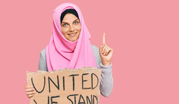 Young caucasian woman holding united we stand banner surprised with an idea or question pointing finger with happy face, number one