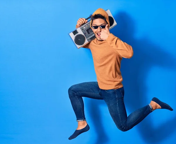 Young handsome latin man smiling happy wearing thug lif sunglasses. Jumping with smile on face holding boombox doing ok sign with thumb up over isolated blue background