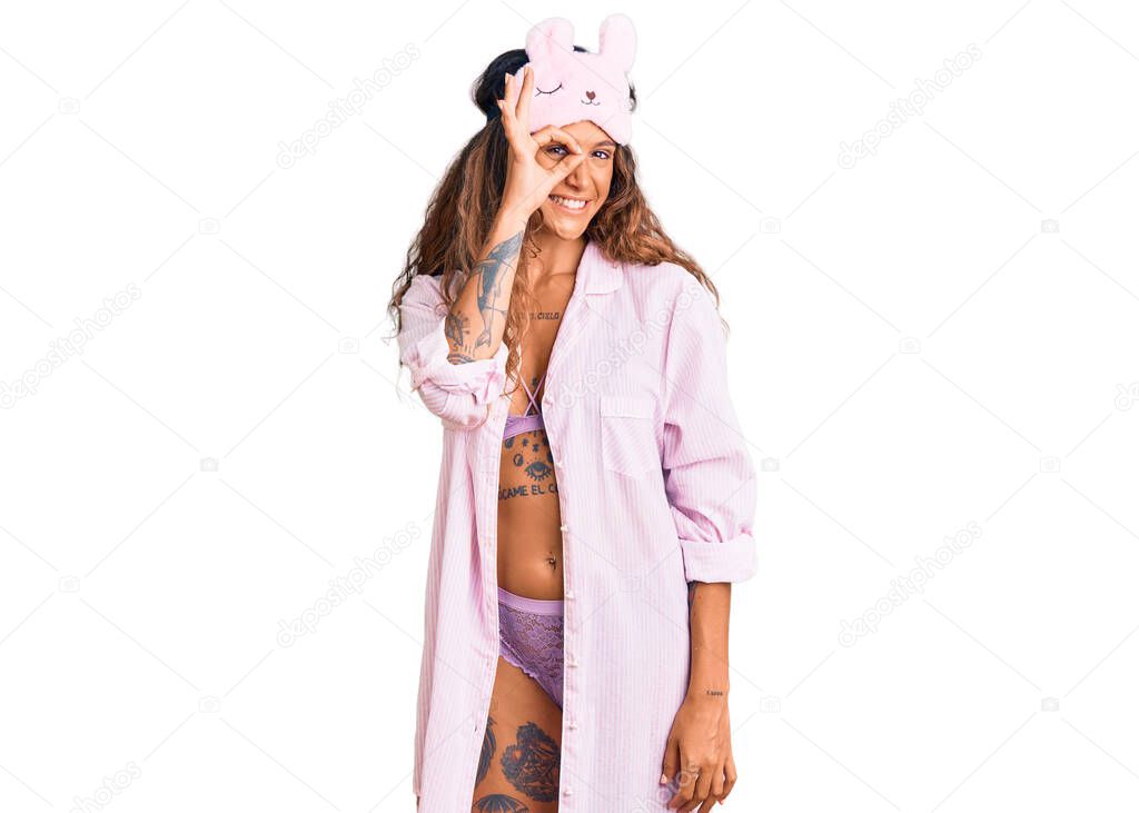 Young hispanic woman with tattoo wearing sleep mask and pajama smiling happy doing ok sign with hand on eye looking through fingers 