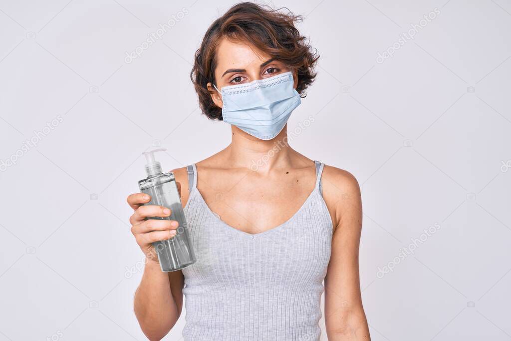 Young hispanic woman wearing medical mask holding hand sanitizer gel thinking attitude and sober expression looking self confident 