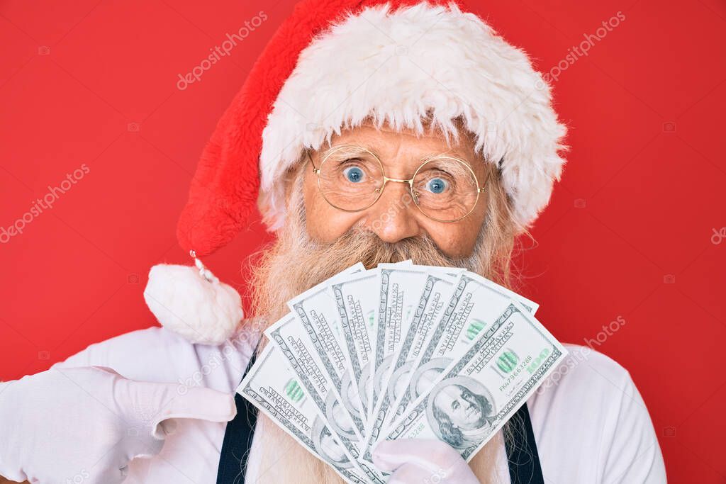 Old senior man with grey hair and long beard wearing santa claus costume holding dollars smiling happy pointing with hand and finger 