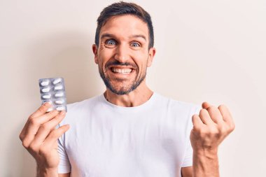 Elinde ilaç hapları olan yakışıklı bir genç beyaz arka planda gururla bağırıyor zaferi ve başarıyı kutluyor.