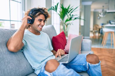 Evde dizüstü bilgisayar ve kulaklık kullanarak mutlu gülümseyen genç İspanyol adam.