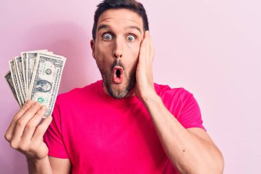 Soyutlanmış pembe arka planda bir deste banknot tutan yakışıklı genç bir adam korkmuş ve ağzı açık şaşkınlık içinde, inançsız bir yüz.