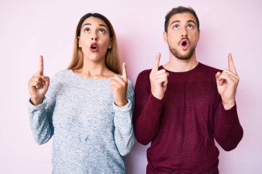 Gündelik giysiler giymiş güzel bir çift şaşırmış ve şaşırmış bir şekilde yukarı bakıp parmaklarını göstererek kollarını kaldırmış.. 