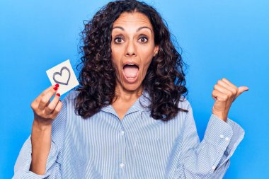 Orta yaşlı, güzel bir kadın, elinde kalp şeklinde bir hatırlatıcı, baş parmağını yukarı kaldırmış, ağzı açık, mutlu bir şekilde gülümsüyor. 