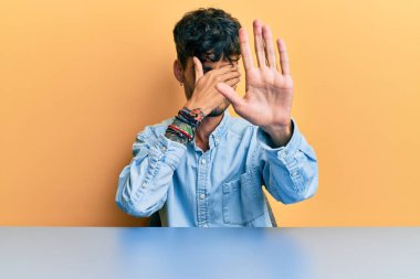 Latin asıllı genç bir adam, gündelik giysiler içinde masada oturuyor, elleriyle gözlerini kapatıyor ve üzgün ve korku ifadeleriyle jestleri durduruyor. utangaç ve olumsuz kavram. 
