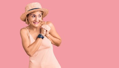 Yazlık şapka takan genç sarışın kadın gergin ve heyecanlı elleri çenesinde yan tarafa bakıyor. 