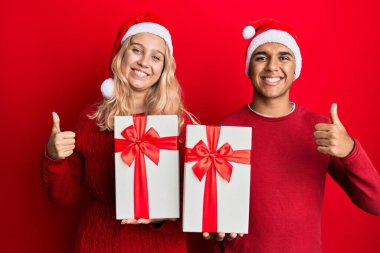Irklar arası genç bir çift noel şapkası takıyor ve ellerinde bir hediye var mutlu ve pozitif gülümsüyorlar, başparmakları havada mükemmel ve onay işareti yapıyorlar. 