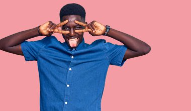 Genç Afro-Amerikan adam günlük kıyafetler giyiyor barış sembolü yapıyor. Yüzünde parmakları var, gülümsüyor ve zaferini gösteriyor. 