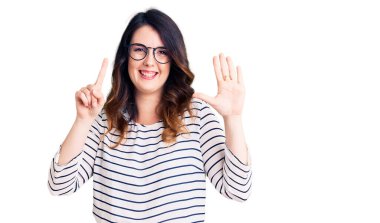 Güzel, genç, esmer bir kadın günlük kıyafetler ve gözlük takıyor. Altıncı parmaklarıyla gösteriyor ve işaret ediyor. Kendinden emin ve mutlu bir şekilde gülümsüyor.. 