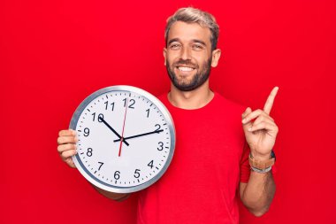 Sakallı, genç, yakışıklı, geri sayım yapan, kırmızı arka planda büyük bir saat kullanan, mutlu, eli ve parmağı yana dönük sarışın bir adam.