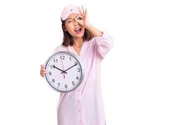 Küçük güzel Çinli kız uyku maskesi ve pijama giymiş elinde büyük bir saat tutarak mutlu mesut gülümsüyor. 