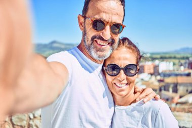 Orta yaşlı, güzel bir çift, günlük kıyafetler ve mutlu bir şekilde gülümseyen güneş gözlüğü takıyor. Yüzünde tebessümle kameranın önünde selfie çekerek kasaba sokaklarında sarılmak..
