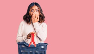 Genç, güzel, melez bir kadın elinde süpermarket alışveriş sepetiyle ağzını eliyle kapatıyor, şok geçiriyor ve hata yapmaktan korkuyor. şaşırmış ifade 