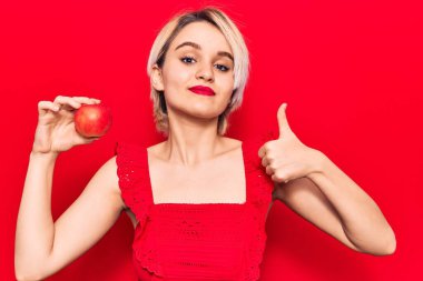 Genç, güzel, sarışın, elinde kırmızı elmayla mutlu ve pozitif gülümseyen, başparmağıyla mükemmel ve onay işareti yapan bir kadın. 