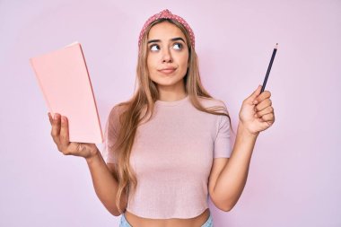 Genç, güzel, sarışın bir kadın elinde kitap ve kalemle gülümsüyor, yan tarafa bakıyor ve düşünüyor.. 