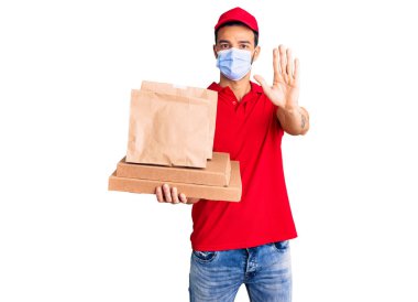 Yakışıklı, İspanyol asıllı genç bir adam yemek dağıtıyor. Covid-19 güvenlik maskesi takıyor ve elinde dur işareti var. Ciddi ve kendinden emin bir ifadeyle. 