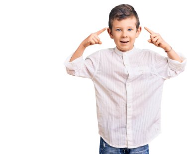 Zarif tişört giyen sevimli sarışın çocuk gülümseyerek iki eliyle başını işaret ediyor, harika bir fikir ya da düşünce, iyi bir hafıza 