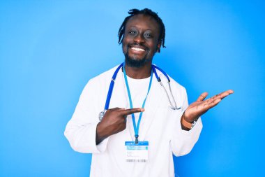 Doktor steteskop ve kimlik kartı takan genç Afrikalı Amerikalı adam kameraya hayret içinde bakıp gülümserken eliyle ve parmağıyla işaret ediyor.. 