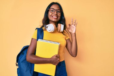 Sırt çantası ve kitap tutan genç Hintli kız parmaklarıyla imza atıp gülümsüyor ve mükemmel bir sembol oluşturuyor. 
