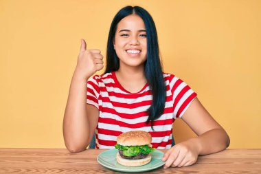 Lezzetli bir klasik burger yiyen genç Asyalı kız mutlu ve pozitif gülümsüyor, başparmağıyla mükemmel ve onay işareti yapıyor. 