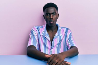 Genç Afro-Amerikan adam, gündelik giysiler giymiş masada oturmuş dudaklarıyla balık suratı yapıyor, çılgınca ve komik bir jest. Komik ifade. 