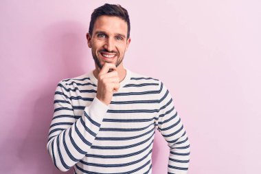 İzole edilmiş pembe arka planda duran sıradan çizgili süveter giyen genç ve yakışıklı bir adam kameraya kollarını kavuşturup, çenesine dokunarak gülümsüyor. Olumlu düşünüyorum..