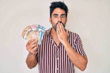 Sakallı yakışıklı İspanyol bir adam Kanada doları tutuyor. Ağzı ağızla örtüyor. Şok olmuş ve hata yapmaktan korkuyor. şaşırmış ifade 
