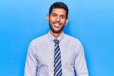 İş kıyafetleri giyen genç Latin adam pozitif ve mutlu görünüyor. Kendine güvenen bir gülümsemeyle ayakta duruyor ve dişlerini gösteriyor. 