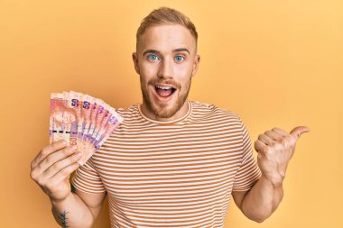 Güney Afrika 'nın 50 rand banknotunu elinde tutan beyaz bir adam başparmağıyla yan tarafa bakıyor ve ağzı açık gülümsüyor. 