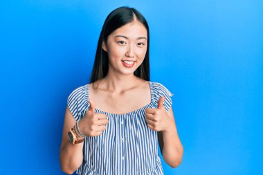 Sıradan çizgili tişört giyen genç Çinli bir kadın. Başarı işareti ile el hareketi yapıyor. Gülümsüyor ve mutlu. Neşeli ifade ve kazanan jest. 